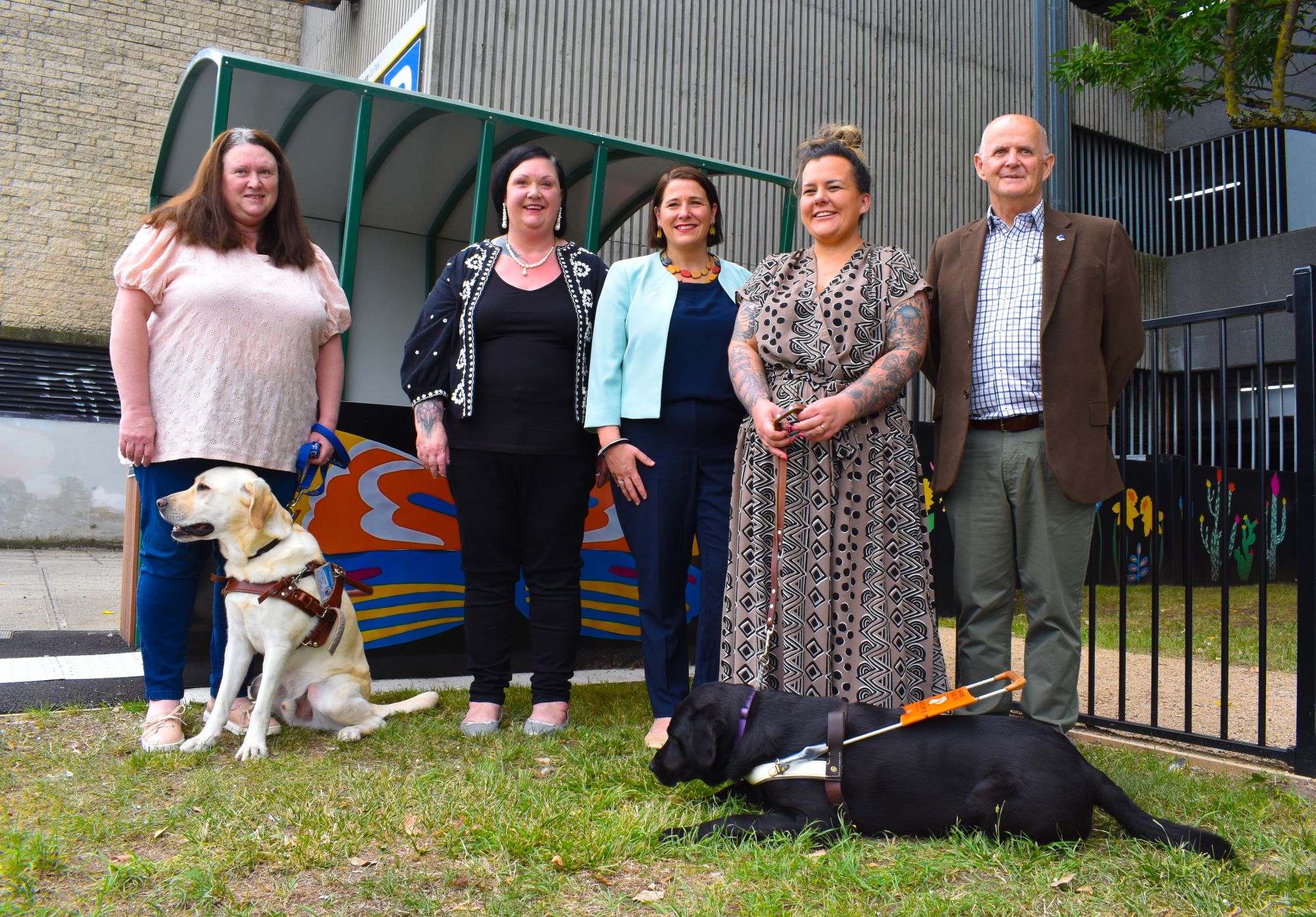 Four women and one man stand in front of a shelter and fenced area, with two assistance dogs. Perf