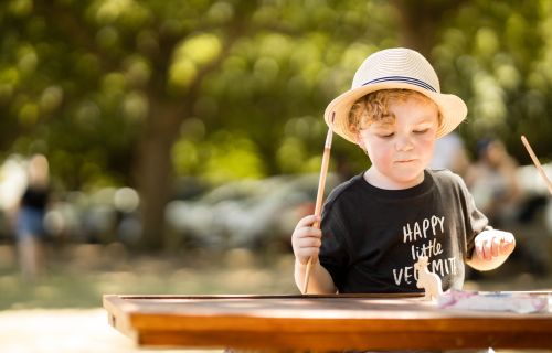 A small child holding paintbrushes at a picnic table