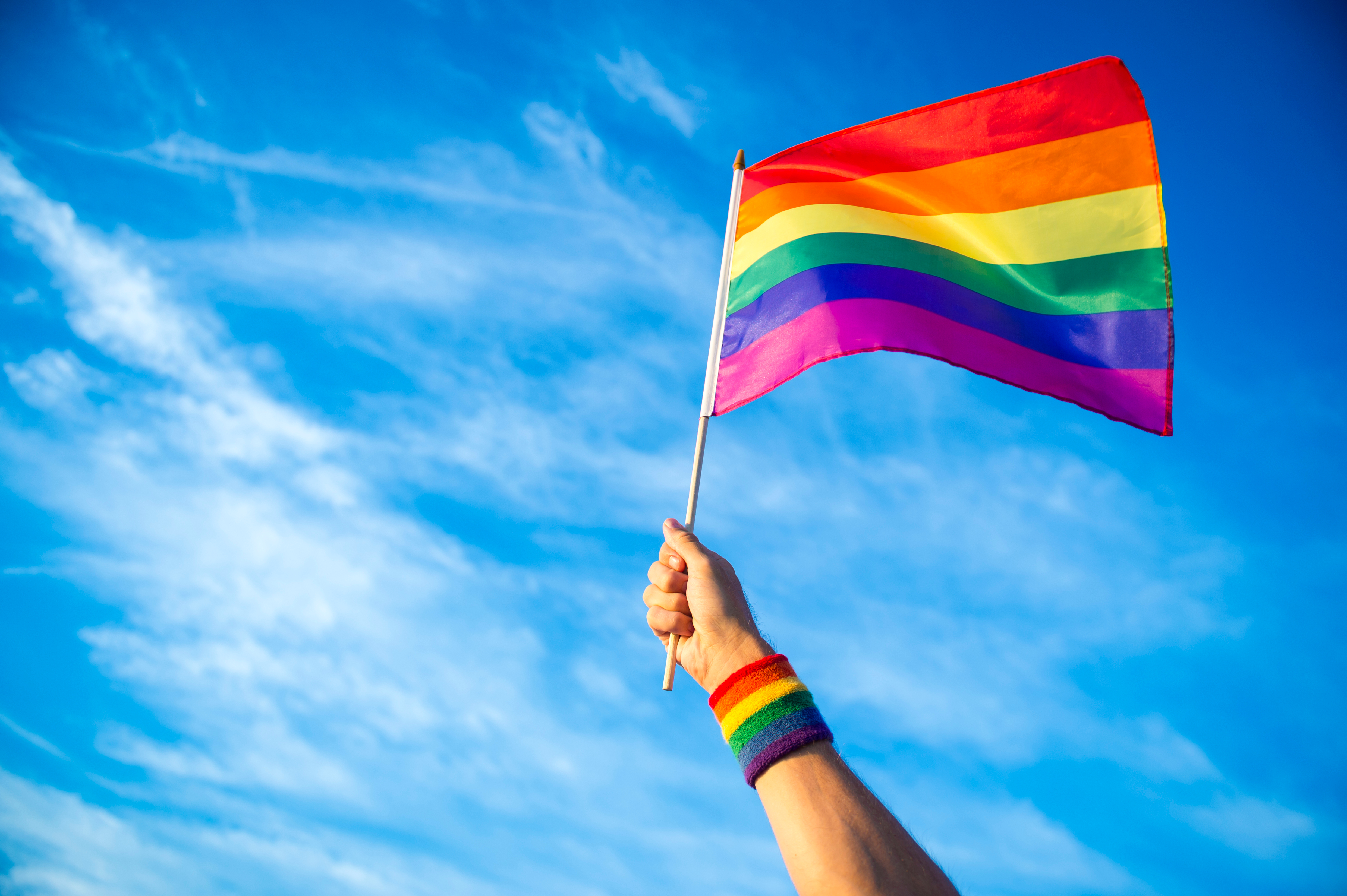 A hand with a rainbow wristband holds a rainbow flag against a blue sky