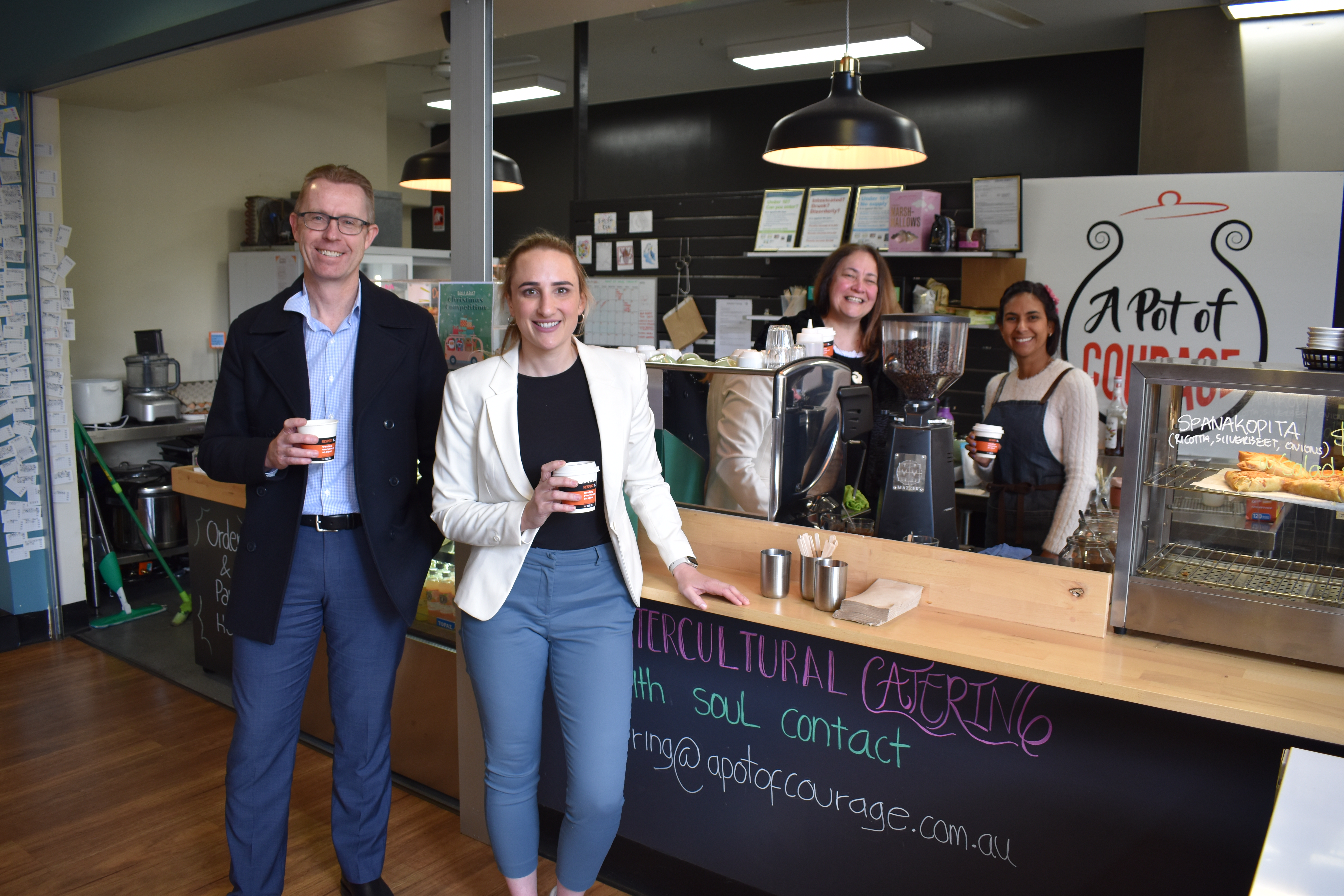 City of Ballarat Chief Executive Officer Evan King and City of Ballarat Deputy Mayor Cr Amy Johnson stand in front of a coffee machine holding coffee cups with promotional sleeves on them, while Pot of Courage founder Shiree Pilkinton stands behind the counter with a Pot of Courage barista. 