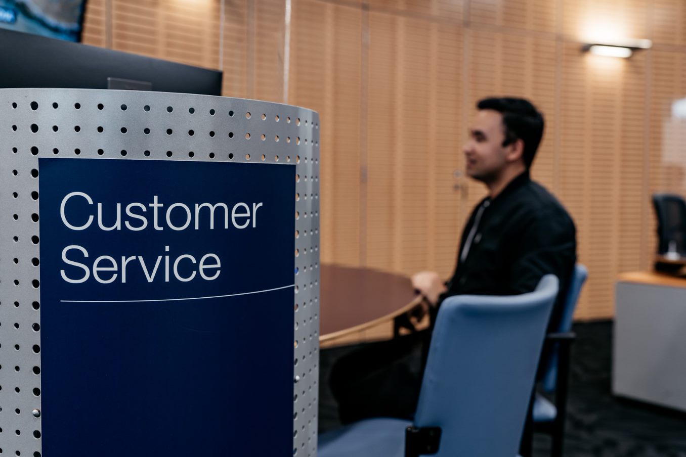 In the foreground, there is a dark blue sign with 'Customer Service' written on it. In the background is a man with sitting at a desk with a blue chair next to him. 