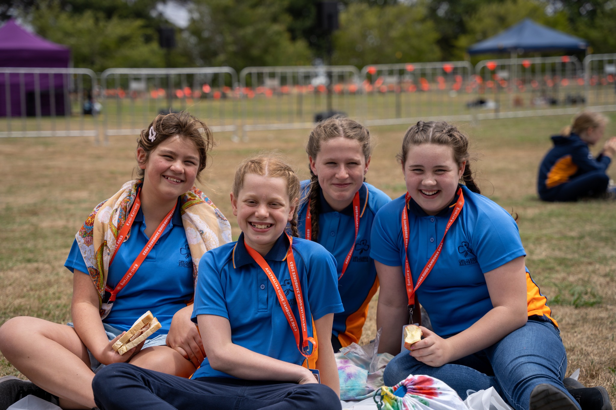 Four girls in blue shirts and orange lanyards sit on a grassed area. 