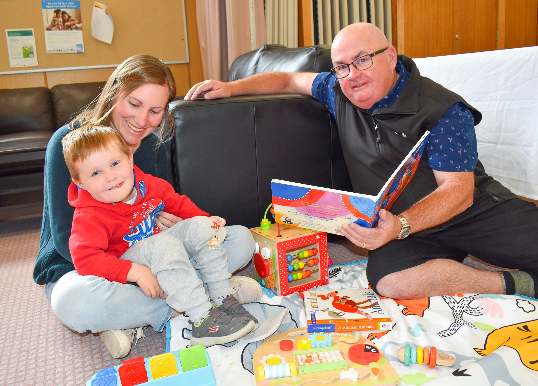 On the left, a young boy sits in a woman's lap and on the right, City of Ballarat Mayor Cr Des Hudson is seated on the ground and holds an open picture storybook. 