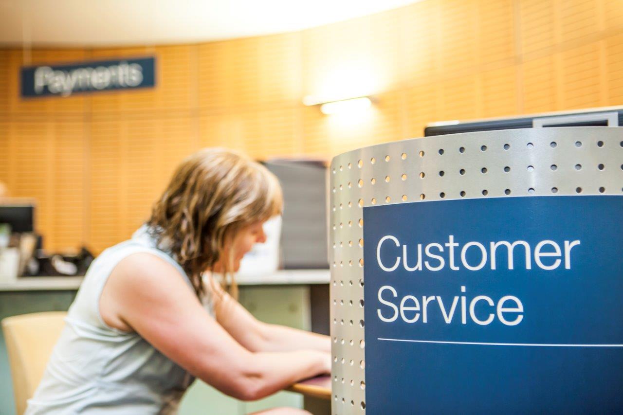 A blue sign saying 'Customer Service' is in the foreground with a seated woman leaning against a desk in the background. 