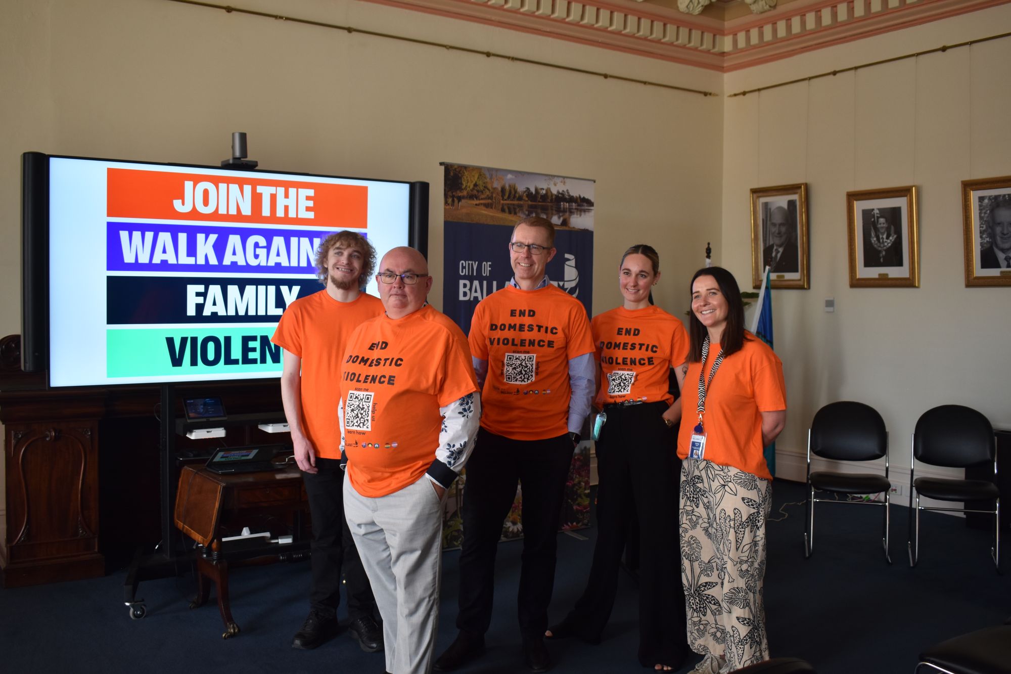 Three men and two women wearing bright orange T-shirts stand in front of a screen that says 'Join the Walk Against Family Violence'