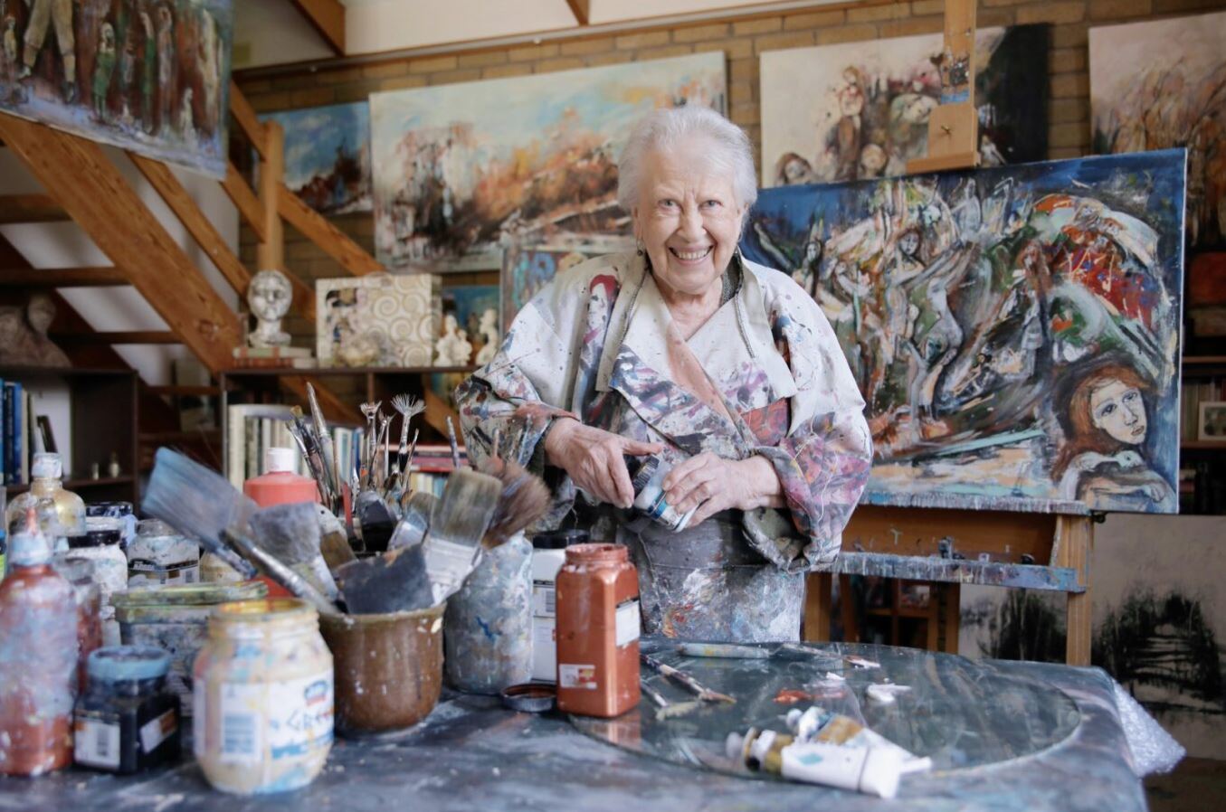 A ninety-two-year-old woman stands in behind a table of paints and brushes, behind her are several colourful pieces of art on canvas. 