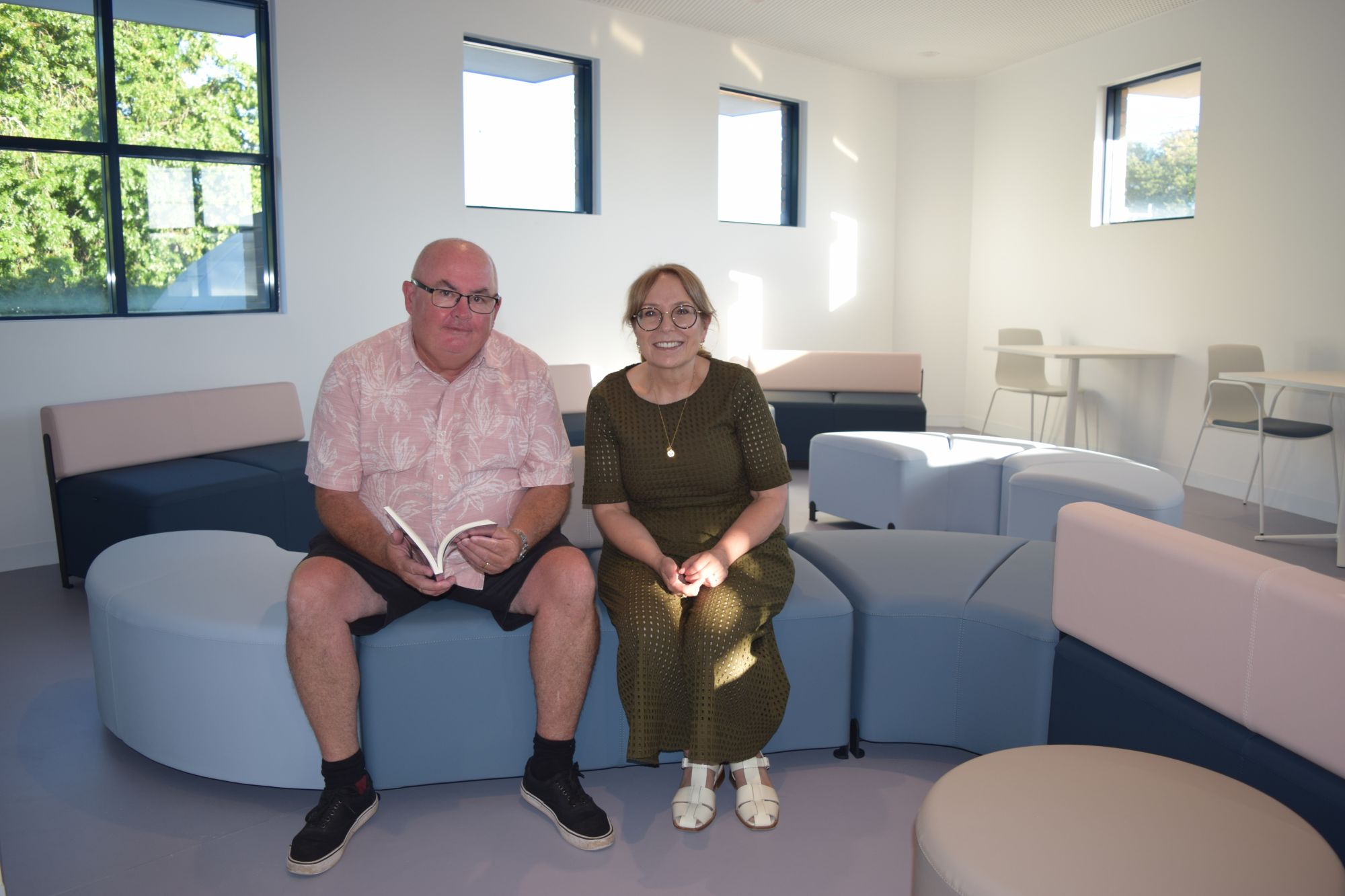 A man and woman sit next to each other in a light-filled room on a blue lounge. The man holds a book. 