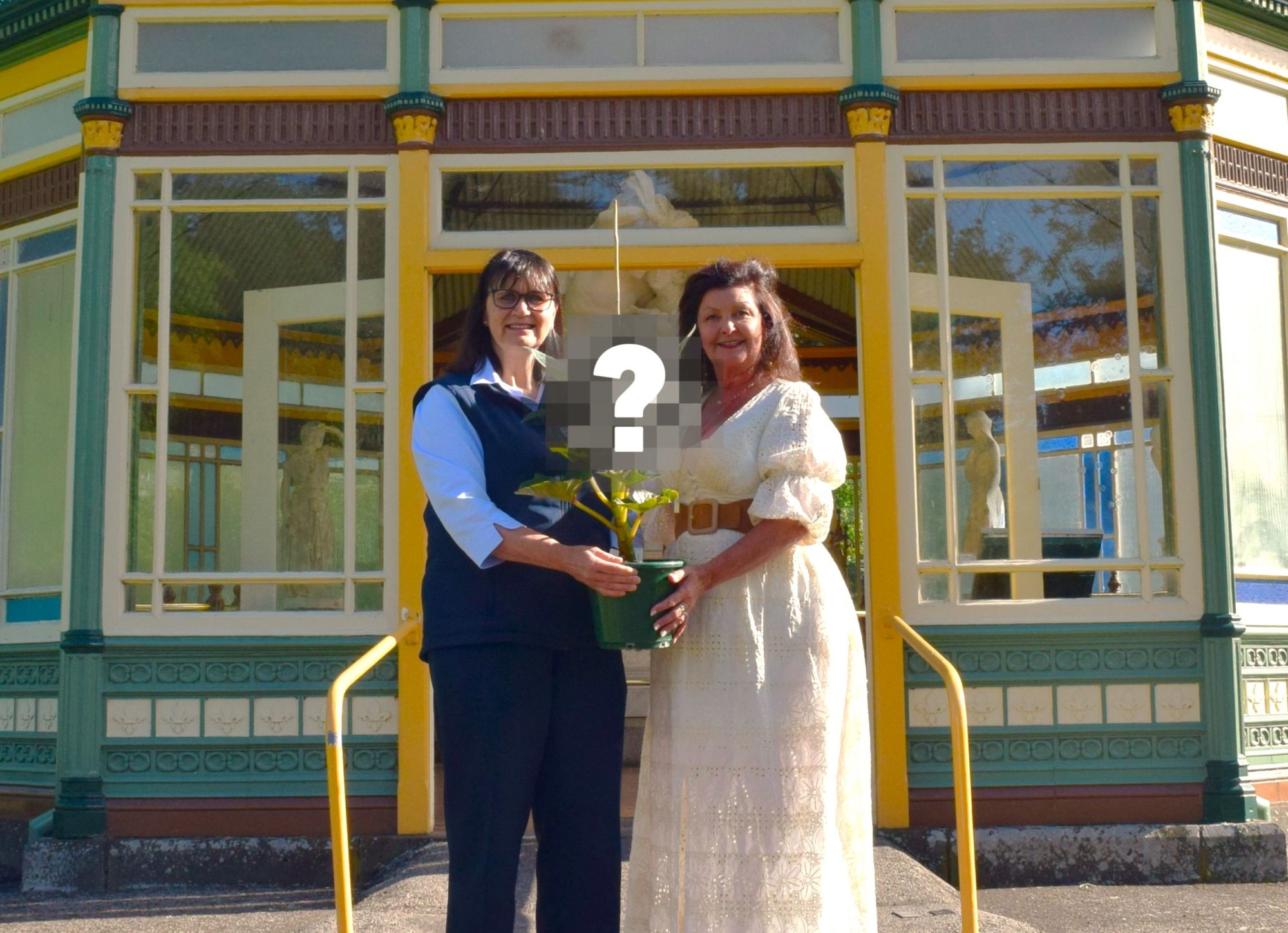 Two women stand in front of a rotunda holding a plant between them. The top of the plant has a question mark over it. 