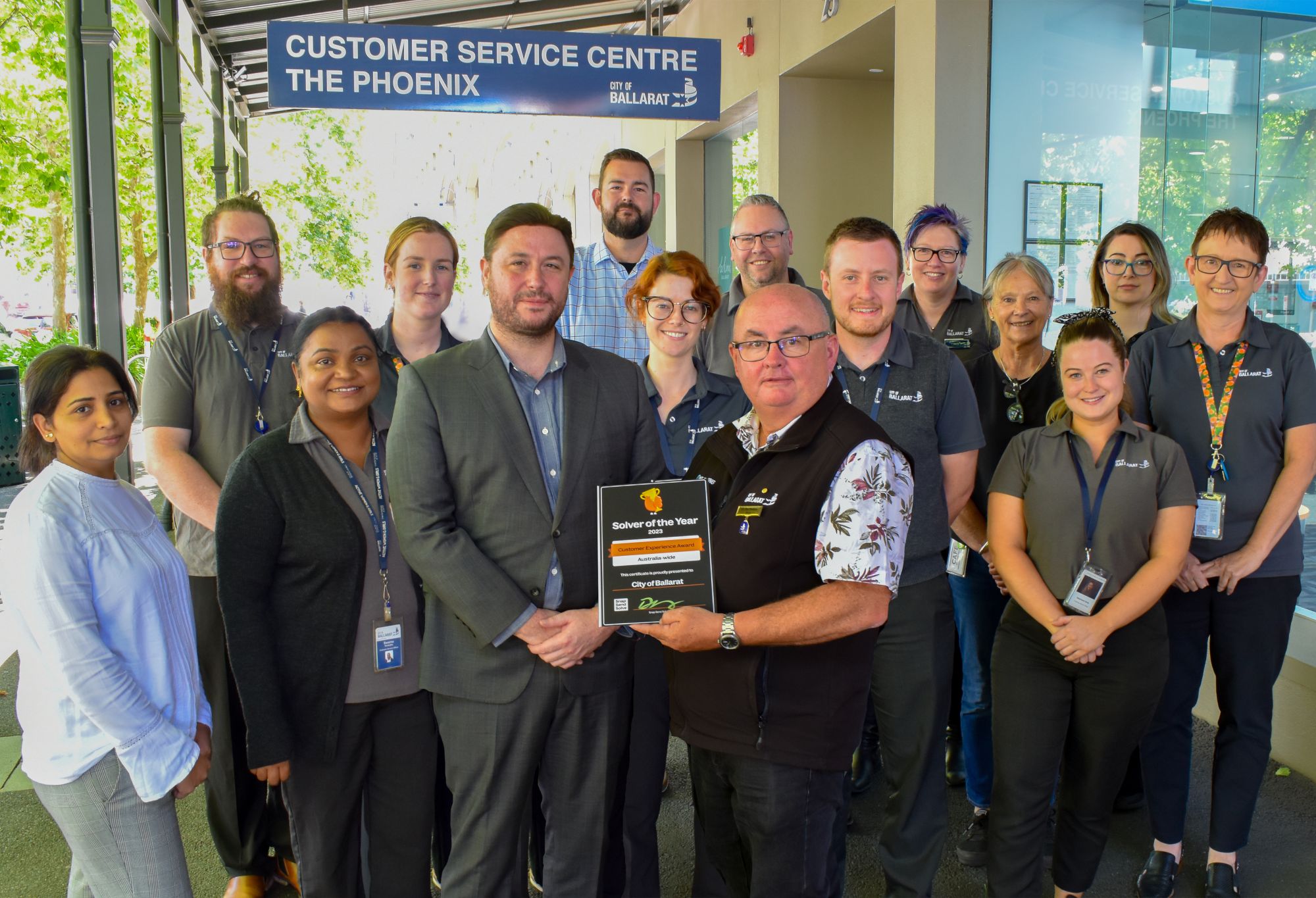 A group of people stand in front of the City of Ballarat Customer Service Centre. The front two people are holding a framed certificate. 