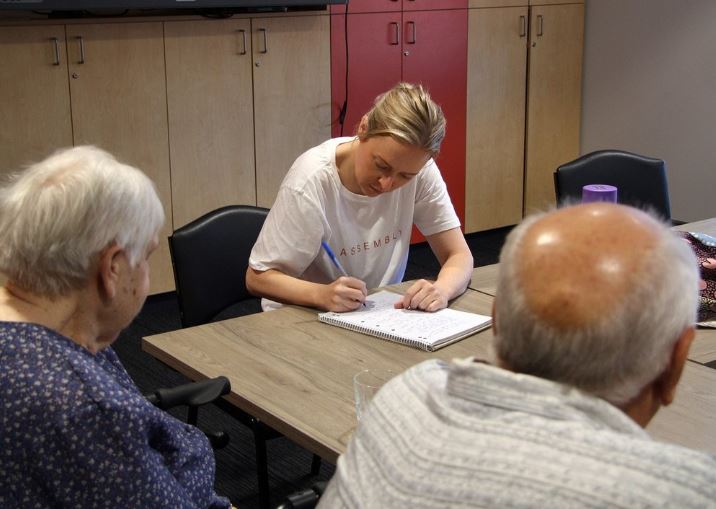 A younger woman writes on a piece of paper across the table from an older woman and man, who are facing away from the camera. 