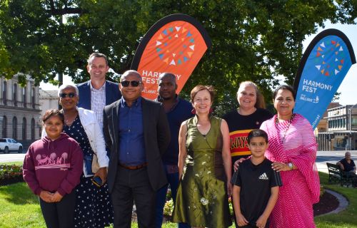 Group of people including Mayor Cr Daniel Moloney and Cr Coates along with Intercultural ambassadors in front of Harmony Fest banners