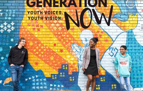 Three diverse young people leaning against a mural reading "generation now"