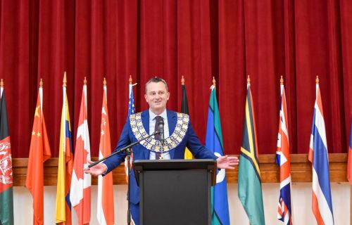 Current City of Ballarat Mayor Cr Daniel Moloney is pictured wearing the Ballarat Mayoral Chains in front of a red curtain and a row of flags.