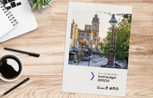 A document with the title 'City of Ballarat Draft Budget 23/24' sits on a table next to a laptop, notebook, cup of black coffee, some pens and a small succulent plant. 
