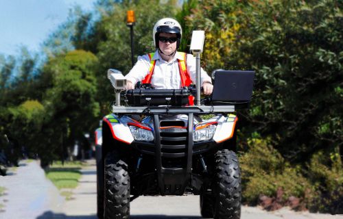 A man a in a helmet drives a large quad bike on a tree-lined footpath. The quad bike has a laptop and camera equipment attached to the front. 