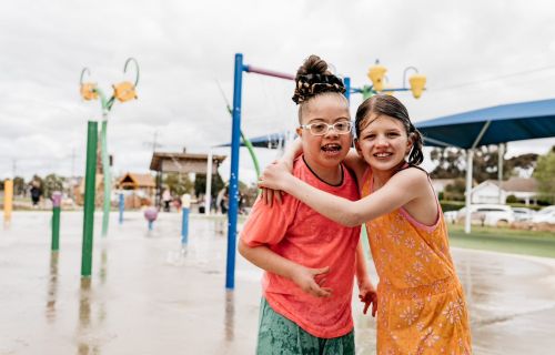 A girl in an orange jumpsuit (left) hugs a girl in a red shirt (right) with splash park equipment in the background. 
