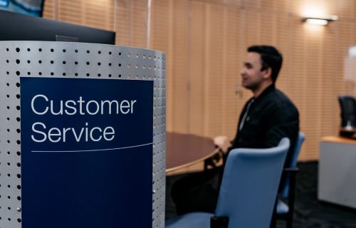 In the foreground, there is a dark blue sign with 'Customer Service' written on it. In the background is a man with sitting at a desk with a blue chair next to him. 