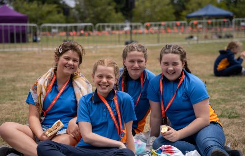 Four girls in blue shirts and orange lanyards sit on a grassed area. 