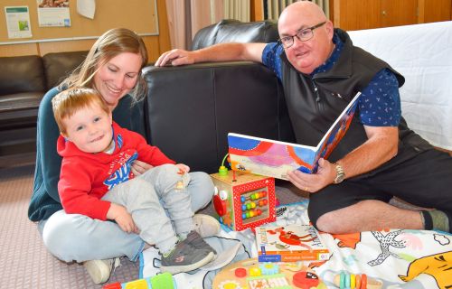 On the left, a young boy sits in a woman's lap and on the right, City of Ballarat Mayor Cr Des Hudson is seated on the ground and holds an open picture storybook. 