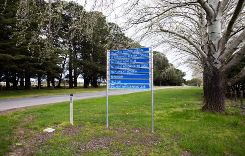 A blue sign with road names on it sits on a lawn in front of a road, with trees behind it. 