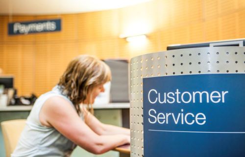 A blue sign saying 'Customer Service' is in the foreground with a seated woman leaning against a desk in the background. 