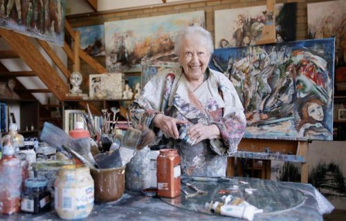 A ninety-two-year-old woman stands in behind a table of paints and brushes, behind her are several colourful pieces of art on canvas. 
