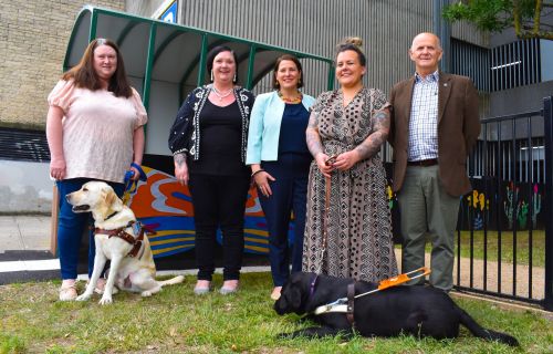 Four women and one man stand in front of a shelter and fenced area, with two assistance dogs. Perf