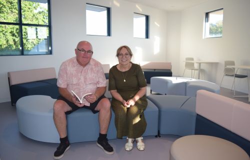 A man and woman sit next to each other in a light-filled room on a blue lounge. The man holds a book. 