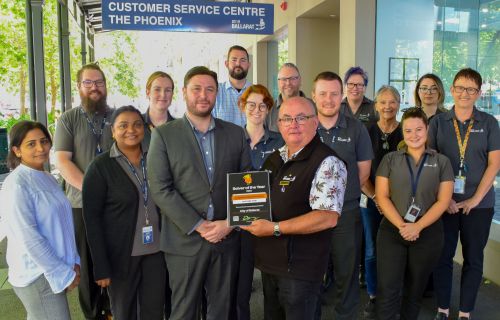 A group of people stand in front of the City of Ballarat Customer Service Centre. The front two people are holding a framed certificate. 