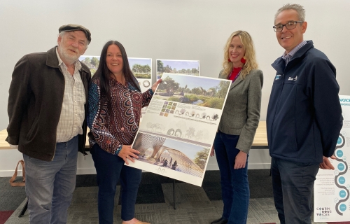Four people stand alongside each other holding a large cardboard display sign with plans for the Continuous Voices Memorial on it. 