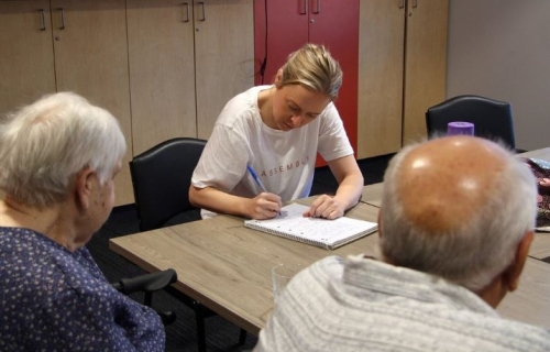 A younger woman writes on a piece of paper across the table from an older woman and man, who are facing away from the camera. 