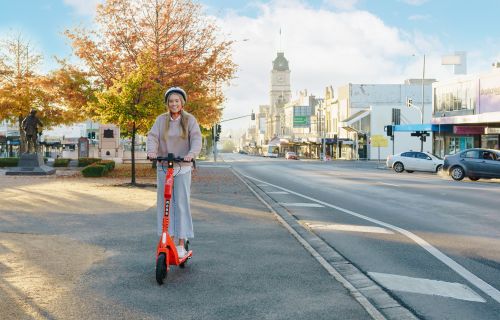 A young woman riding a bright orange e-scooter on Sturt Street with the town hall and Ballarat city in the background