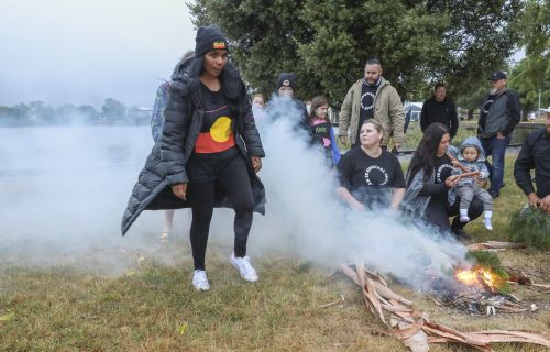 An indigenous woman wearing and aboriginal flag shirt surrounded by the smoke of a fire