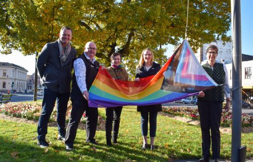 Cr Daniel Moloney, Mayor Des Hudson, Cr Belinda Coates, Cr Amy Johnson and a member of the public hold the Progress Pride flag up. The flag is attached to the flag pole and about to be hoisted up. Everyone is smiling.