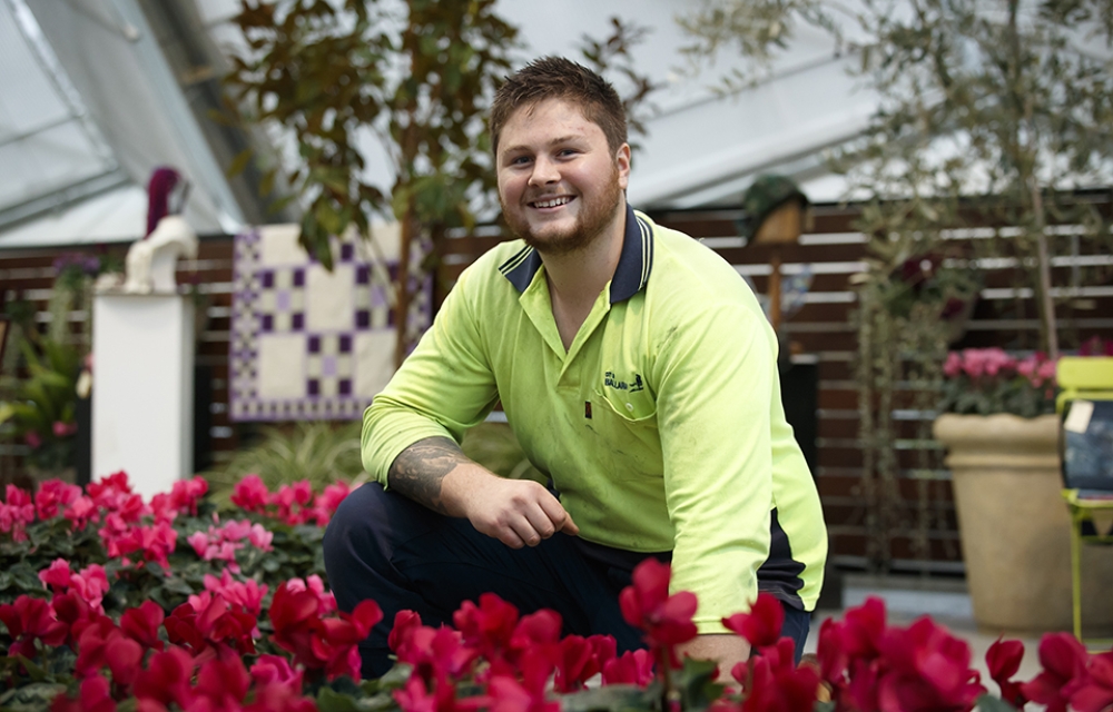 a young man kneeling in a garden bed smiling at the camera