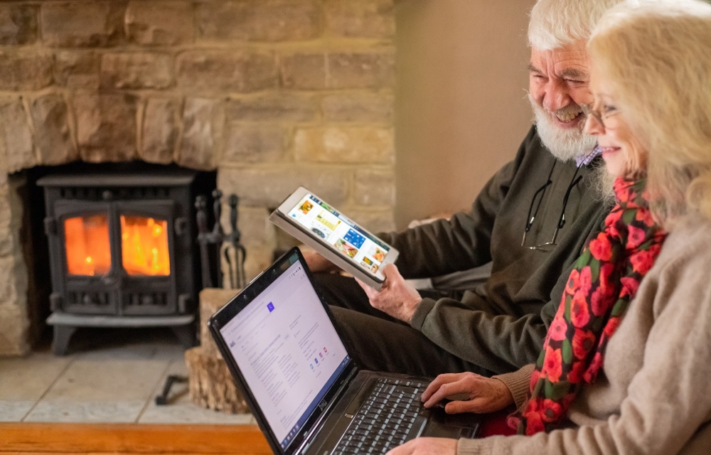 Older man and woman sitting beside each other in a room with a log fire, using a tablet and laptop to explore the internet together.