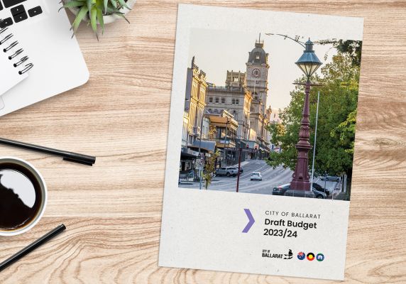 A document with the title 'City of Ballarat Draft Budget 23/24' sits on a table next to a laptop, notebook, cup of black coffee, some pens and a small succulent plant.