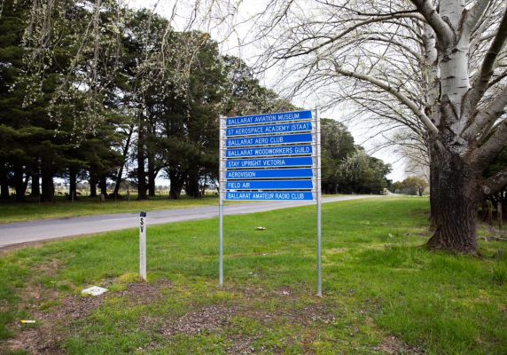 This image shows a blue sign situated on green grass with a road and a row of trees in the background.