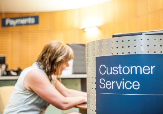 A woman sits next to a blue sign that says 'Customer Service'.