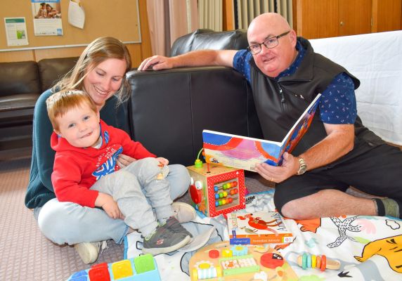 On the left, a young boy sits in a woman's lap and on the right, City of Ballarat Mayor Cr Des Hudson is seated on the ground and holds an open picture storybook.