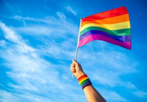 A hand with a rainbow wristband holds a rainbow flag against a blue sky