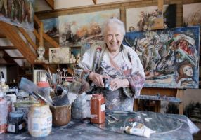 A ninety-two-year-old woman stands in behind a table of paints and brushes, behind her are several colourful pieces of art on canvas. 