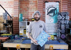 A smiling man stands in front of a workbench holding a colourful piece of art
