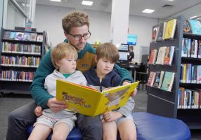 A man and two small children read a book together among the shelves of a library
