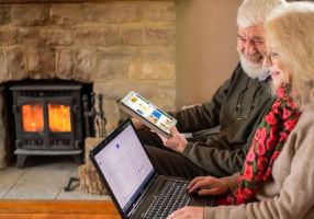 Older man and woman sitting beside each other in a room with a log fire, using a tablet and laptop to explore the internet together. 