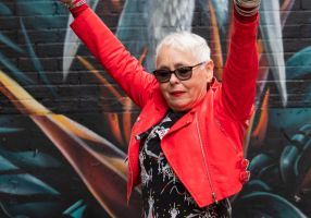 Older woman with short hair, dark glasses and red leather jacket holding her arms up high in front of a mural of a bird.